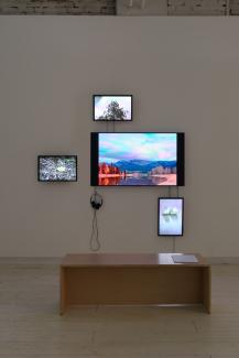 An installation of four TV monitors arranged with one large in the centre, and three small at North, South, West. In front of the monitors is a simple plywood bench.