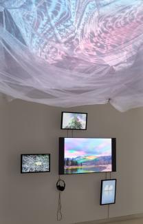 An installation of four TV monitors arranged with one large in the centre, and three small at North, South, West. In the foreground, white netting hangs from above on which pink and blue textures are projected.