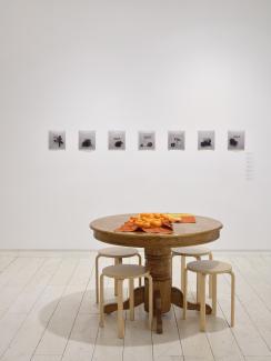 A wooden pedestal table (oak and reminiscent of 90s kitchens) with four wooden stools (birch, modern design) sits in front of a gallery wall. On the table lay two Bojagi (traditional Korean wrapping cloth), and a number of bright orange tangerines. behind the table, is a row of seven small black and white photographs, unframed.