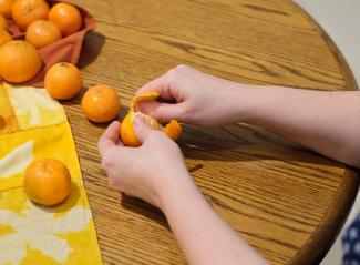 Two hands peeling a tangerine at a wooden table. On the left we see a corner of a bright yellow Bojagi (traditional Korean wrapping cloth), and a number of bright orange tangerines.