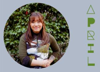 Circular image of head shot of April, wearing a graphic jersey shirt with her arms are her waist, standing against a vibrant leafy bush. The portrait is placed in a dark pale blue grey background with her stylized name in grass green to the right.