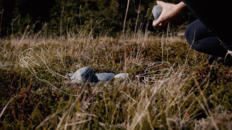 worm's eye view of a person's hands holding a large stone, crouched above a few stones in the grass. The image is overlaid with a white line drawing of hands beading.