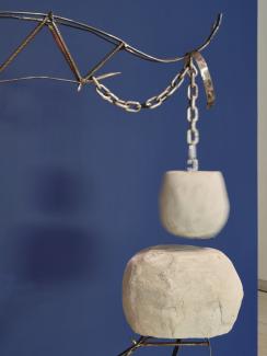 Image of two boulder-like objects suspended from a welded metal arm, against a dark blue background. The upper object is blurred due to its movement, as it hovers above the lower object.