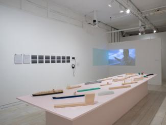 installation image of a gallery exhibition featuring a larger wooden table on which there are models of boat shapes, small photographs in a line on the wall behind it, and a video projection in the background