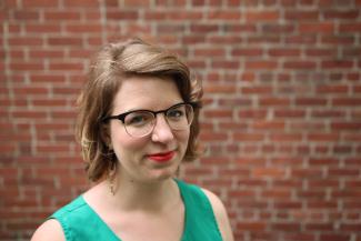 a smiling woman with chin length golden brown hair and horn-rimmed glasses, wearing a green tank top, standing against a brick wall.