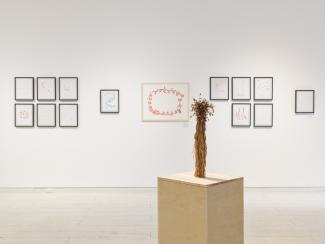 In the foreground a bundle of dried flowers sits upright on a wooden plinth. In the background there is many framed works, mostly weavings, and handmade paper with thread embedded within. 
