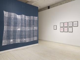 In install shot on the corner of the gallery. The wall to the left is painted blue, with white weaving hanging in front of it. The wall to the right is white, with framed pieces of handmade paper with thread embedded in. 