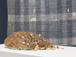 A woven basket made from dried flowers, grass, thread, and paper. It lies on its side on a white table in the foreground. In the background there is a white weaving in front of a blue wall. 
