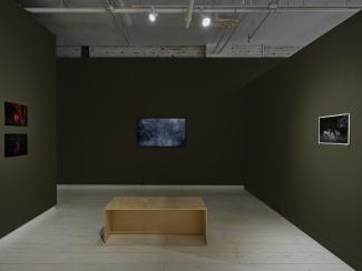 Dark-painted gallery walls feature eight colourful photographs on the left. Nearby, a wooden bench faces a dark-toned video display installation. A small black and white print is located to the right of the video installation.