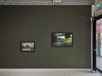 Two photographic works by Steven Cottingham hang in the gallery. The image on the right hangs slightly higher, and shows a simulated forest landscape projected across a dark shooting range. The image on the left shows stacks of computer servers, clad in military green and resting on a wooden pallet. 