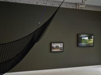 Two photographic works by Steven Cottingham hang in the gallery. The image on the right hangs slightly higher, and shows a simulated forest landscape projected across a dark shooting range. The image on the left shows stacks of computer servers, clad in military green and resting on a wooden pallet. A cargo net cuts across the foreground of the image, suspended from the gallery ceiling.