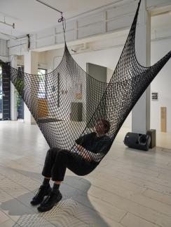 A cargo net, part of Serena Lee's sound installation, is suspended from the gallery ceiling. A person is laying in the net like a hammock, headed tilted back to listen to sound coming from a large speaker on the floor behind them. 