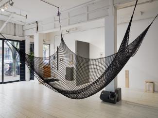 A cargo net, part of Serena Lee's sound installation, is suspended in the centre of the gallery. In the background, letter sized paper sheets line the wall. The papers look like they've come from a desktop printer—one has a drone silhouette, another has a news article, another an academic text.
