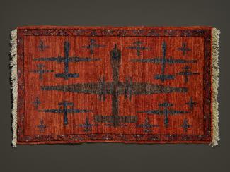 A small, hand-knotted rug by the Weaver hangs on the wall. The background is a rusty red, and a brown predator drone dominates the centre of the image. Four smaller drones flank the central figure in each quadrant, with smaller drones surrounding those. The geometric design and earth tones make the rug appear quite traditional, save for the subject matter.