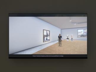 A still image from Steven Cottingham's As Far As The Drone Can See is shown on a monitor. It is a "machinima"—a video filmed within a video game world—from Arma3, showing a feminine character standing within an art gallery. The captions read: "times of national emergency create enormous possibilities for change".