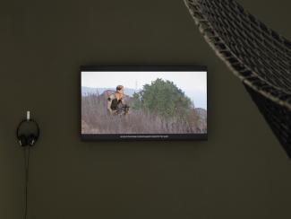 A still image from Steven Cottingham's As Far As The Drone Can See is shown on a monitor. It is a "machinima"—a video filmed within a video game world—from Arma3, showing a feminine figure crouched behind a bush. Captions pick up mid-sentence, reading "... who die in the process of producing genetic material for their queen." 