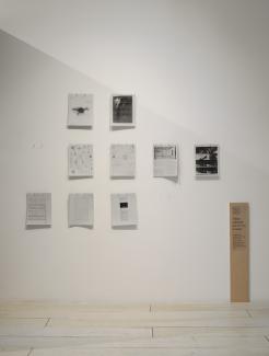 Grey letter sized readings are pinned in a neat grid on a wall. Some have news articles, academic texts, and images of drones. Some spots on the grid are empty where the readers have been taken by visitors. Signage in the gallery accompanies them: "These readings are for you to keep."