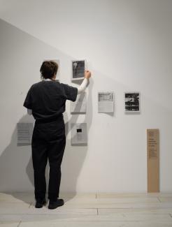 A person pulls a text from the reader wall. The other texts include news articles, academic texts, and images of drones. Some spots on the grid are empty where the readers have been taken by visitors. Signage in the gallery accompanies them: "These readings are for you to keep."