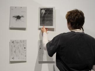 A close up of a person in a black shirt taking a paper from the reader wall. The cover page of the reader is beside them, reading: SMALL GODS: ON AERIAL VISUALITY, RESONANCE, AND THE DRONE.