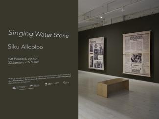 A photograph of the title wall, reading "Singing Water Stone - Siku Allooloo." Two large canvas prints of newspaper enlargements are visible in the background. 
