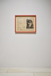 A red-framed newspaper centrefold hangs on a white wall. A photograph of an older woman's face is visible on the righthand spread.