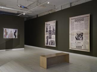Two large canvas prints stretch from floor to ceiling in the gallery. They show enlargements of a newspaper titled Indígena: News From Indian America. There is a pastel-coloured silk print hanging toward the middle of the gallery. 