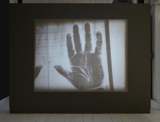 A video projection on a dark wall shows a hand. The footage is in negative, the hand is dark against a light background. 